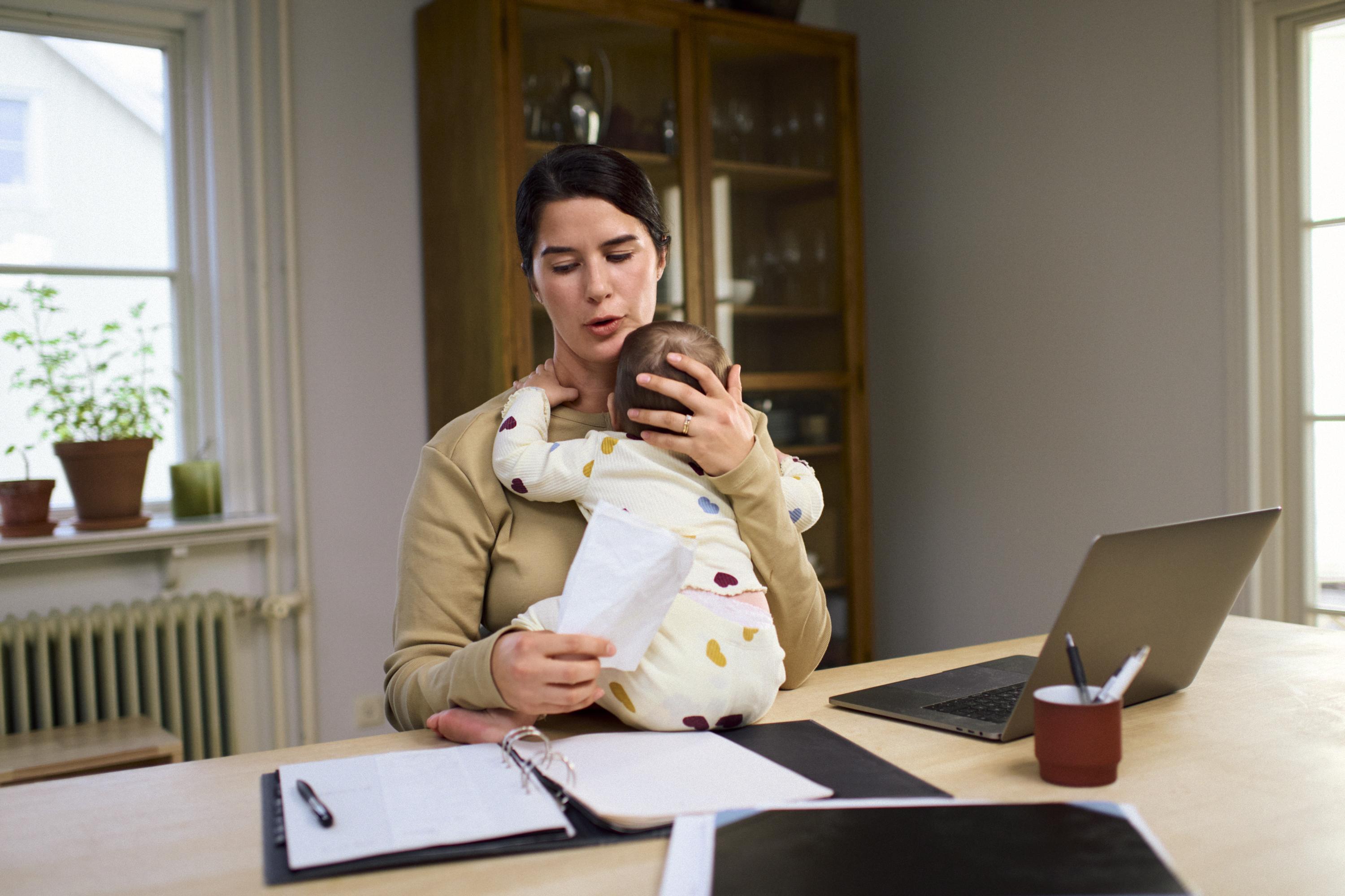 A woman sitting at a table with a laptop and papers while holding a small baby..