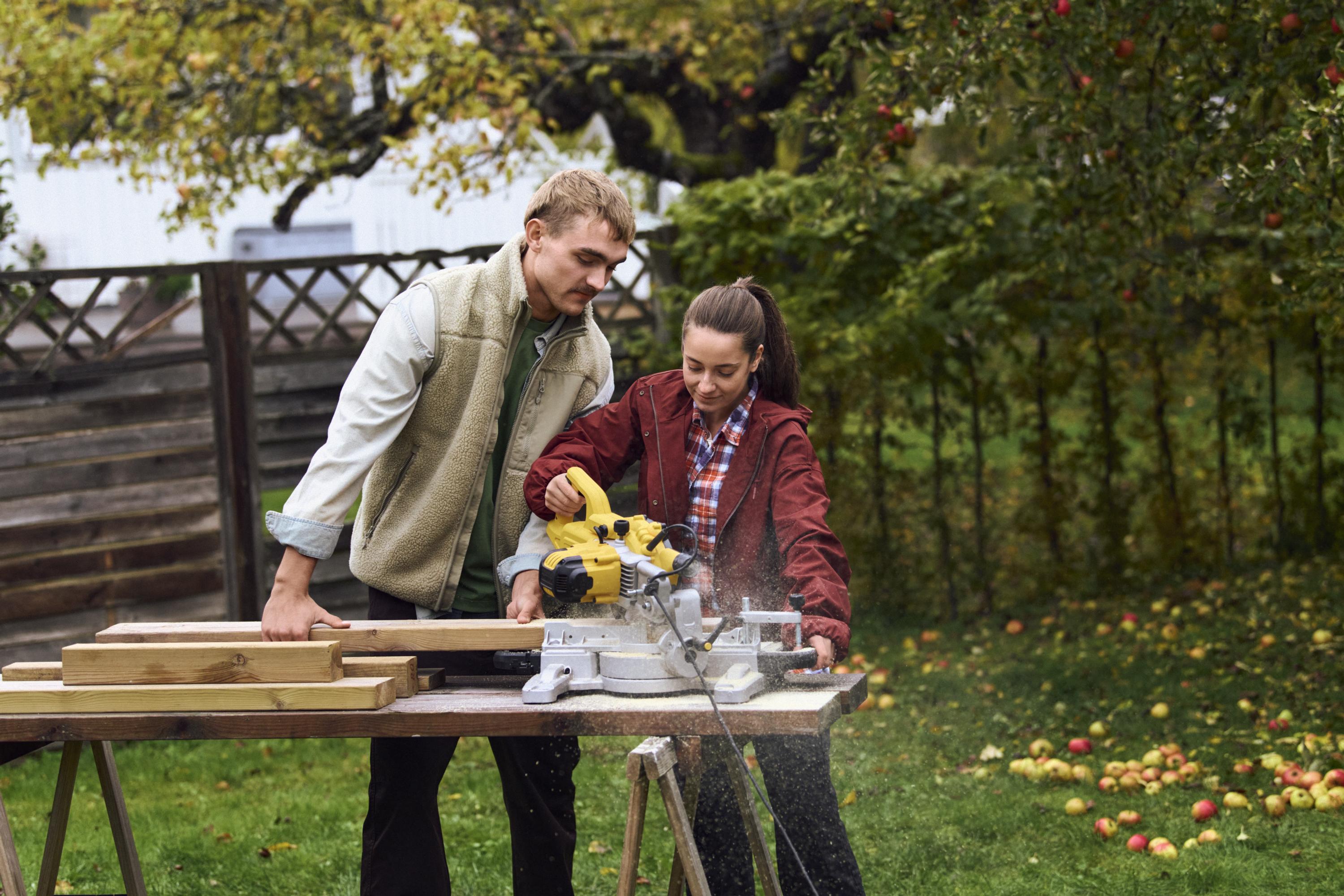 En kvinna och en man sågar brädor med en kapsåg i en trädgård.